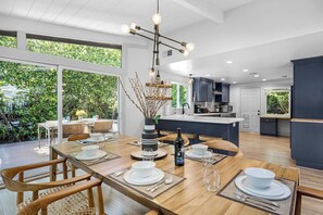 Een moderne keuken met een centraal eiland, grote ramen en een eetgedeelte met een houten tafel voor vier personen.