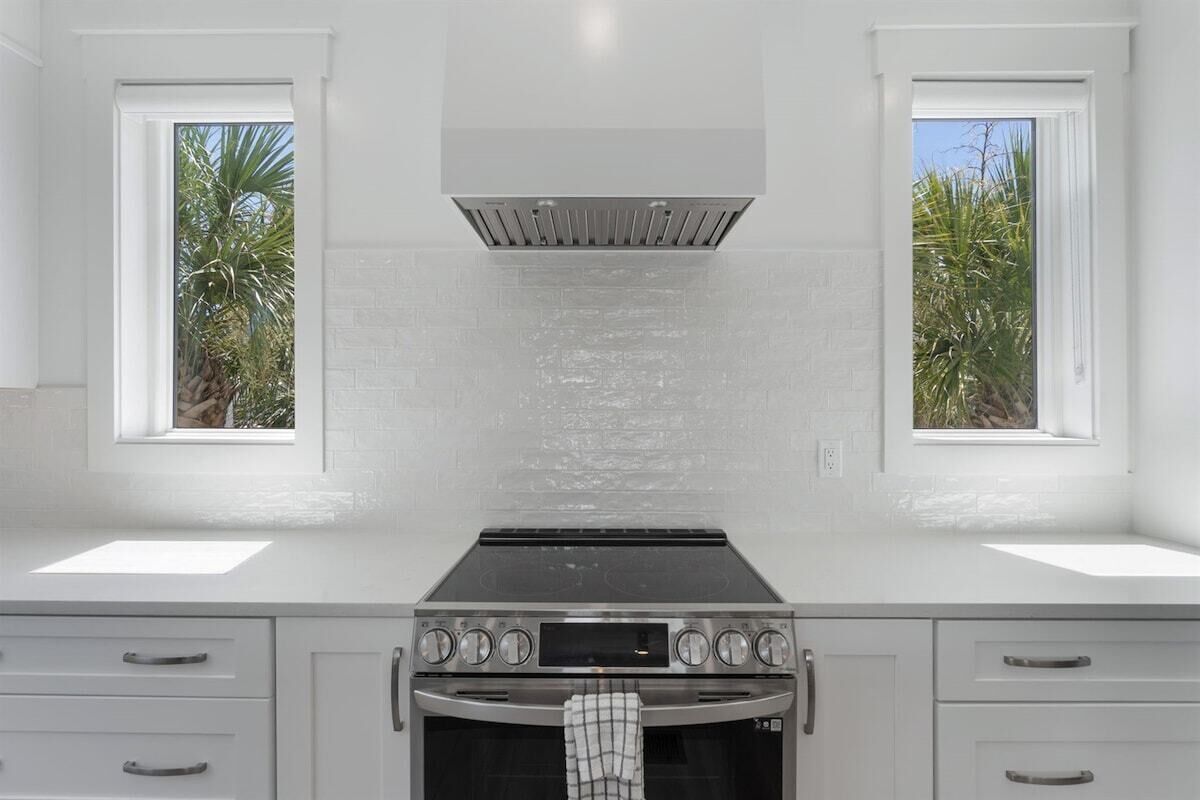 A kitchen with a stainless steel stove, two windows with views of palm trees, and white cabinetry.