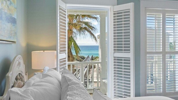 A bedroom with a bed, a nightstand, a lamp, and a view of the ocean through open shutters. - Ocean front: Private Pool + Private Beach Access (Seagrove Beach)