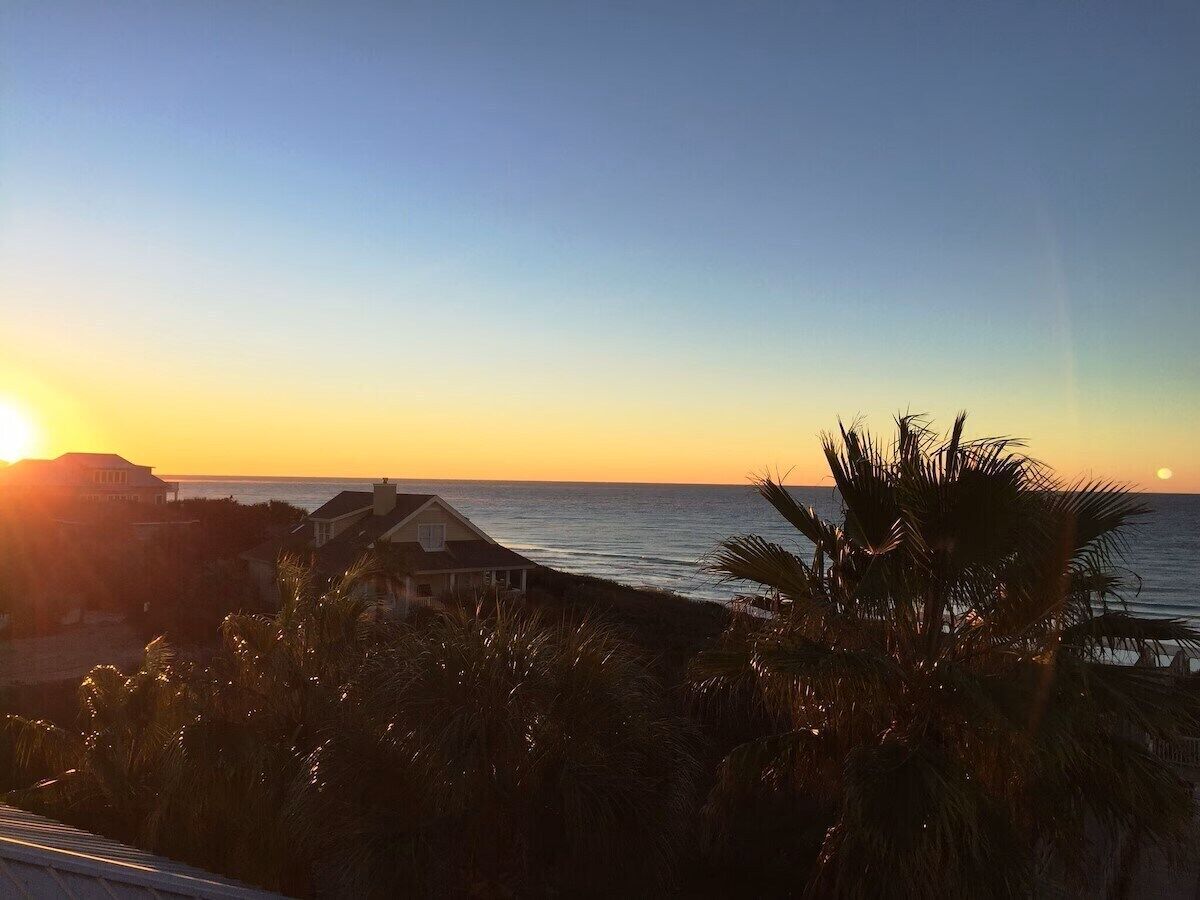 A coastal view with a sunset, palm trees, and a house by the sea.