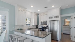 A modern kitchen with white cabinets, a stainless steel refrigerator, a microwave, and a granite countertop. There are four white bar stools at the island. - Ocean front: Private Pool + Private Beach Access (Seagrove Beach)