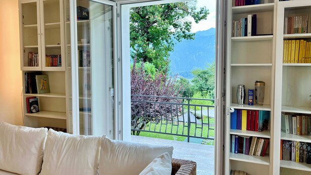 A room with a white sofa, open bookshelves, and a view of greenery and mountains through a glass door.
