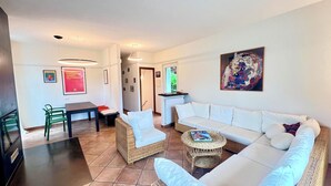 A modern living room with a white sofa, wicker chairs, a dining table, and a small coffee table. There are framed pictures on the walls and a painting above the sofa.