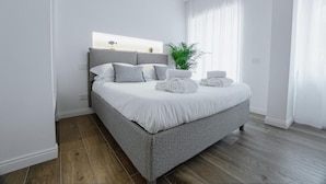 Une chambre moderne avec un lit recouvert d’un tissu gris, du linge de lit blanc et une plante sur la table de chevet.