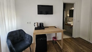 Une chambre d’hôtel compacte comprenant un bureau en bois, une chaise noire, un téléviseur fixé au mur, une machine à café et une petite poubelle.