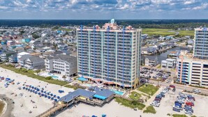 A high-rise hotel with multiple floors, a swimming pool, and a beach area with umbrellas. - Prince Resort Oceanfront 3BR Pools Lazy River (north myrtle beach)