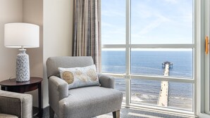 A room with a large window overlooking the sea, a grey armchair with decorative pillows, a lamp on a side table, and a view of a pier. - Prince Resort Oceanfront 3BR Pools Lazy River (north myrtle beach)