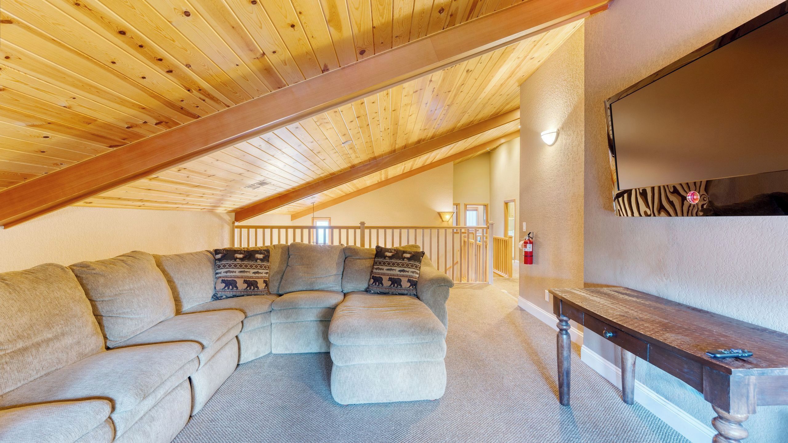 Une chambre à coucher confortable, aménagée en style loft, avec un plafond en bois, un canapé d’angle, un petit pouf, un banc en bois et un téléviseur fixé au mur.