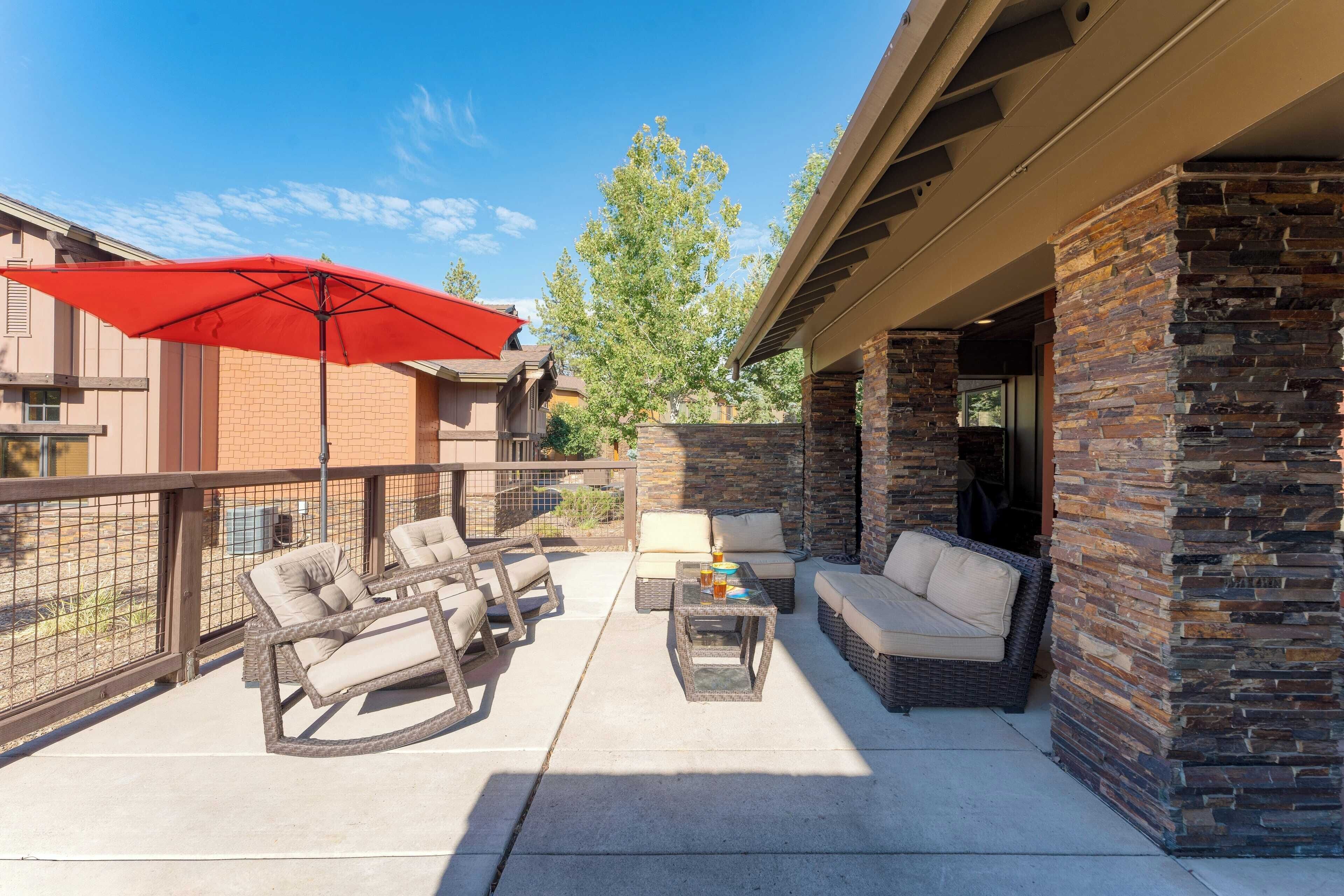An outdoor patio with wicker furniture, a red umbrella, and a stone wall.