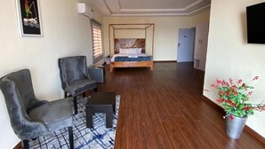 Una habitación de hotel con una cama, dos sillas, una mesita, una planta en maceta y un cuadro en la pared.