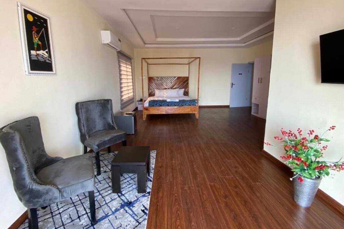 A hotel room with a bed, two chairs, a small table, a potted plant, and a painting on the wall.