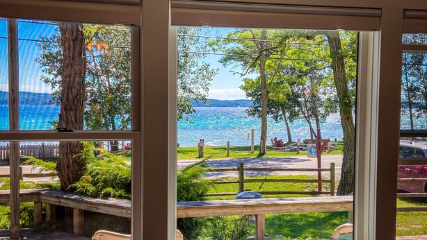 Une vue depuis une fenêtre donnant sur un espace au bord du lac, avec une terrasse en bois, une table de pique-nique et des chaises.