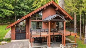 A modern two-story cabin with a stone foundation, large windows, and a balcony with outdoor seating. - Charming 4-bedroom Ski house in Stratton with Shuttle or walk to slope. (Stratton)