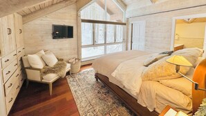 A bedroom with a sloped ceiling, wooden walls, a large bed, a chair, a television, and a window with a view of trees. - Charming 4-bedroom Ski house in Stratton with Shuttle or walk to slope. (Stratton)