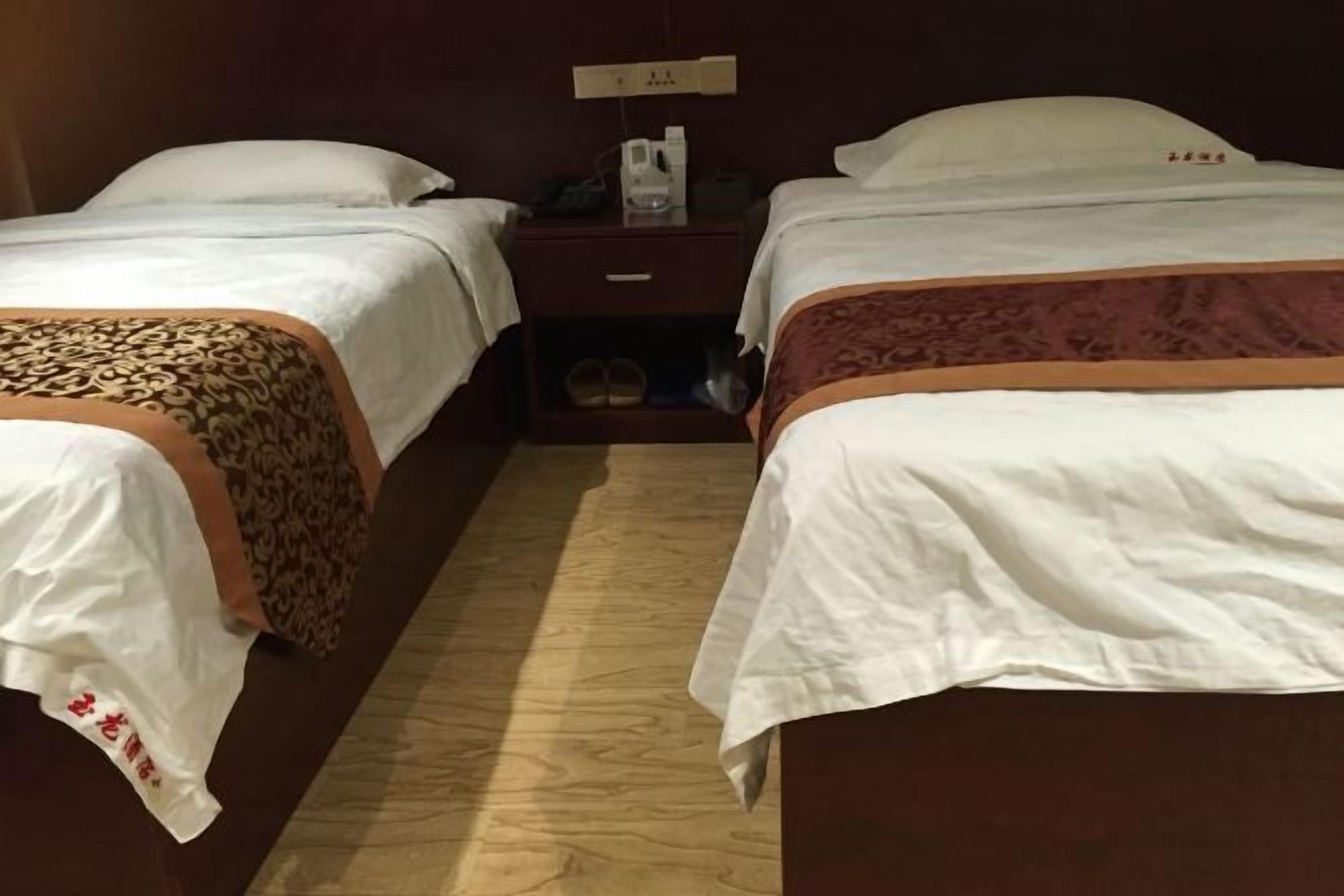 A hotel room with two beds, each with a white bedspread and a brown decorative border. There is a nightstand between the beds with a phone and a small lamp.