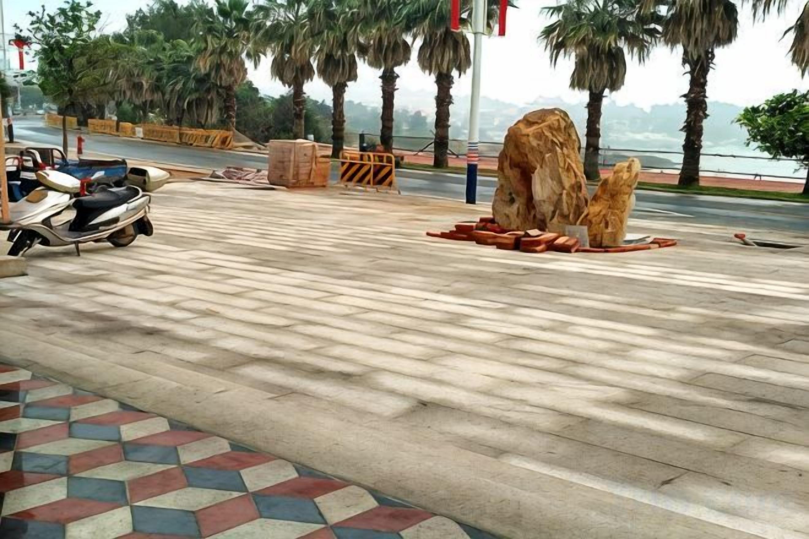 A paved area with a scooter parked, a large rock sculpture, and palm trees in the background.