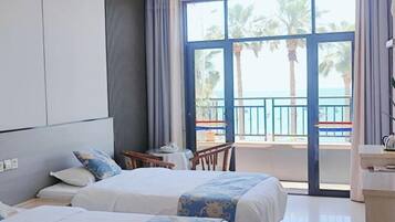A hotel room with two beds, a balcony with a view of palm trees and the sea, and a small table with a chair.
