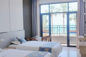 Ein Hotelzimmer mit zwei Betten, einem Balkon mit Blick auf Palmen und das Meer sowie einem kleinen Tisch mit einem Stuhl.