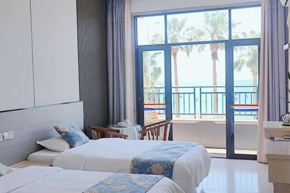 Habitación de hotel con dos camas, un balcón que ofrece vistas a palmeras y al mar, y una pequeña mesa con una silla.