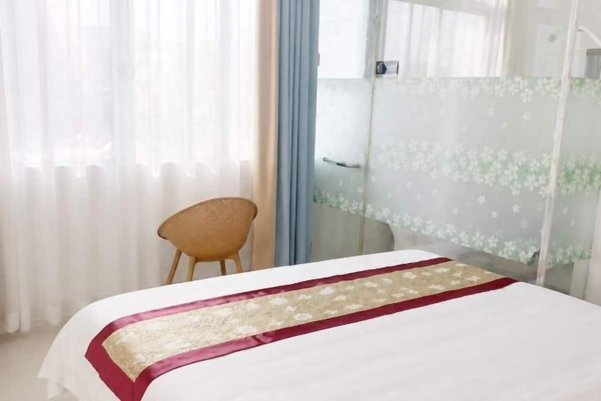 Hotellihuone, jossa on sänky, tuoli, lasiseinä ja ikkuna, jossa on läpikuultavat verhot.