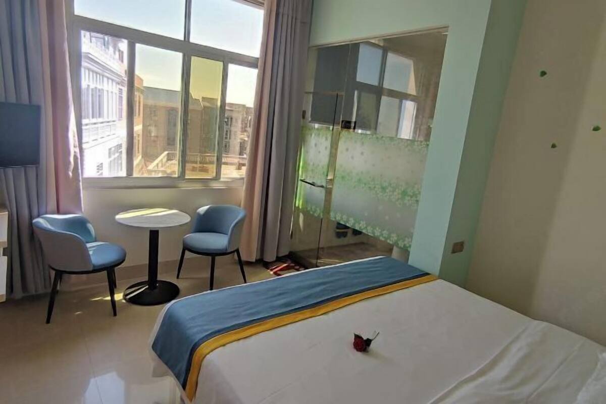 Hotellihuone, jossa on sänky, pieni pöytä, kaksi tuolia, ikkuna, josta on näkymä rakennuksiin, ja pieni keittotila.