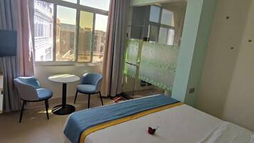 A hotel room with a bed, a small table, two chairs, a window with a view of buildings, and a kitchenette area.