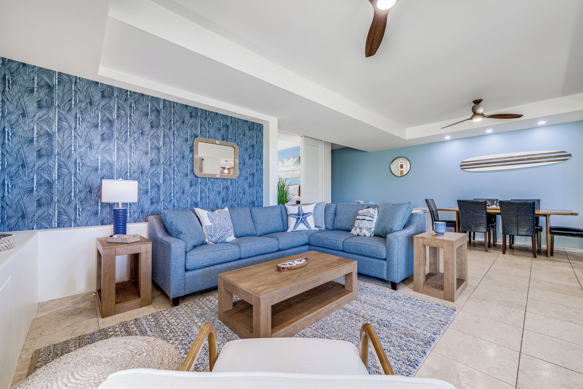 Un salón moderno con papel tapiz tropical azul, un sofá modular, una mesa de centro de madera y un comedor con decoración de tabla de surf.