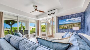 A modern living room with a large blue sofa, a ceiling fan, and a view of a tropical landscape through large sliding doors. - Gorgeous Renovated Condo Walking Distance to Beaches!! Large Private Lanai (Kihei)