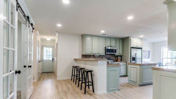 A spacious kitchen with light wood flooring, a central island with stools, and light green cabinetry. - The Main Street Cottage: Dog-Friendly, Central DFW location, Walk to Main St (Lewisville)