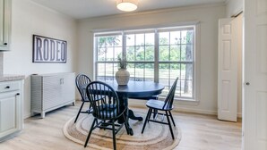 A dining area with a round table, four chairs, a vase, and a large window with a view of greenery. - The Main Street Cottage: Dog-Friendly, Central DFW location, Walk to Main St (Lewisville)