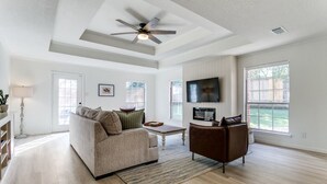 A spacious living room with a ceiling fan, a fireplace, a television, and large windows. - The Main Street Cottage: Dog-Friendly, Central DFW location, Walk to Main St (Lewisville)