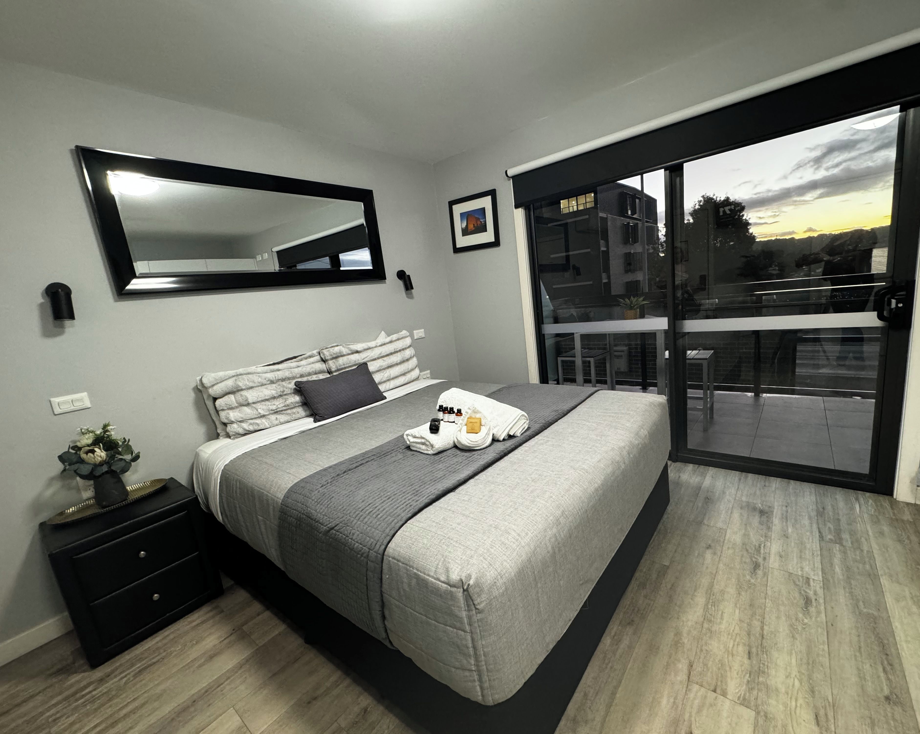 A modern bedroom with a large bed, a bedside table, a mirror, and a sliding glass door leading to a balcony.