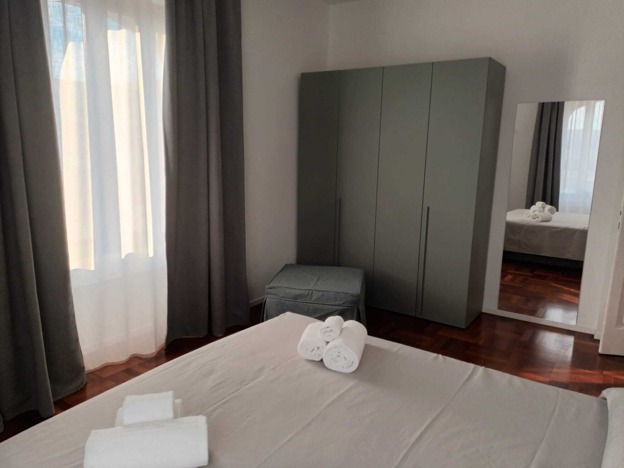 Une chambre d’hôtel avec un lit, une armoire grise, un petit banc et un miroir.