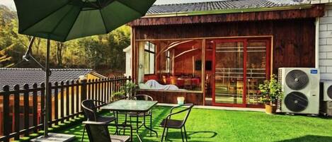 An outdoor patio with a table and chairs, a green umbrella, and a wooden building with a red door.