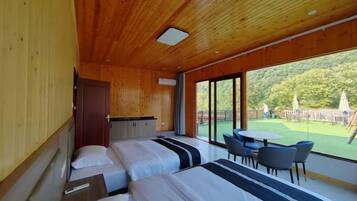Una habitación moderna con paredes y techo de madera, dos camas con ropa de cama a rayas, una mesita pequeña y un amplio ventanal corredera que da a un espacio exterior con césped y sombrillas.