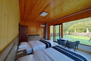 Una habitación moderna con paredes y techo de madera, dos camas con ropa de cama a rayas, una mesita pequeña y un amplio ventanal corredera que da a un espacio exterior con césped y sombrillas.