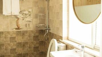 Un baño con ducha, inodoro y lavabo. Se observa un perchero con toallas y un florero con flores en la pared.
