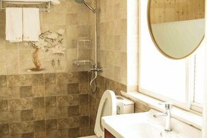 Un baño con ducha, inodoro y lavabo. Se observa un perchero con toallas y un florero con flores en la pared.