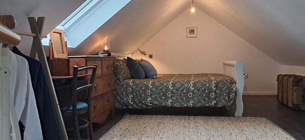 A cozy attic bedroom with a sloped ceiling, a wooden dresser, a chair, a bed with floral bedding, and a suitcase. - The Stables - Charming cottage in beautiful West Cork (Skibbereen)