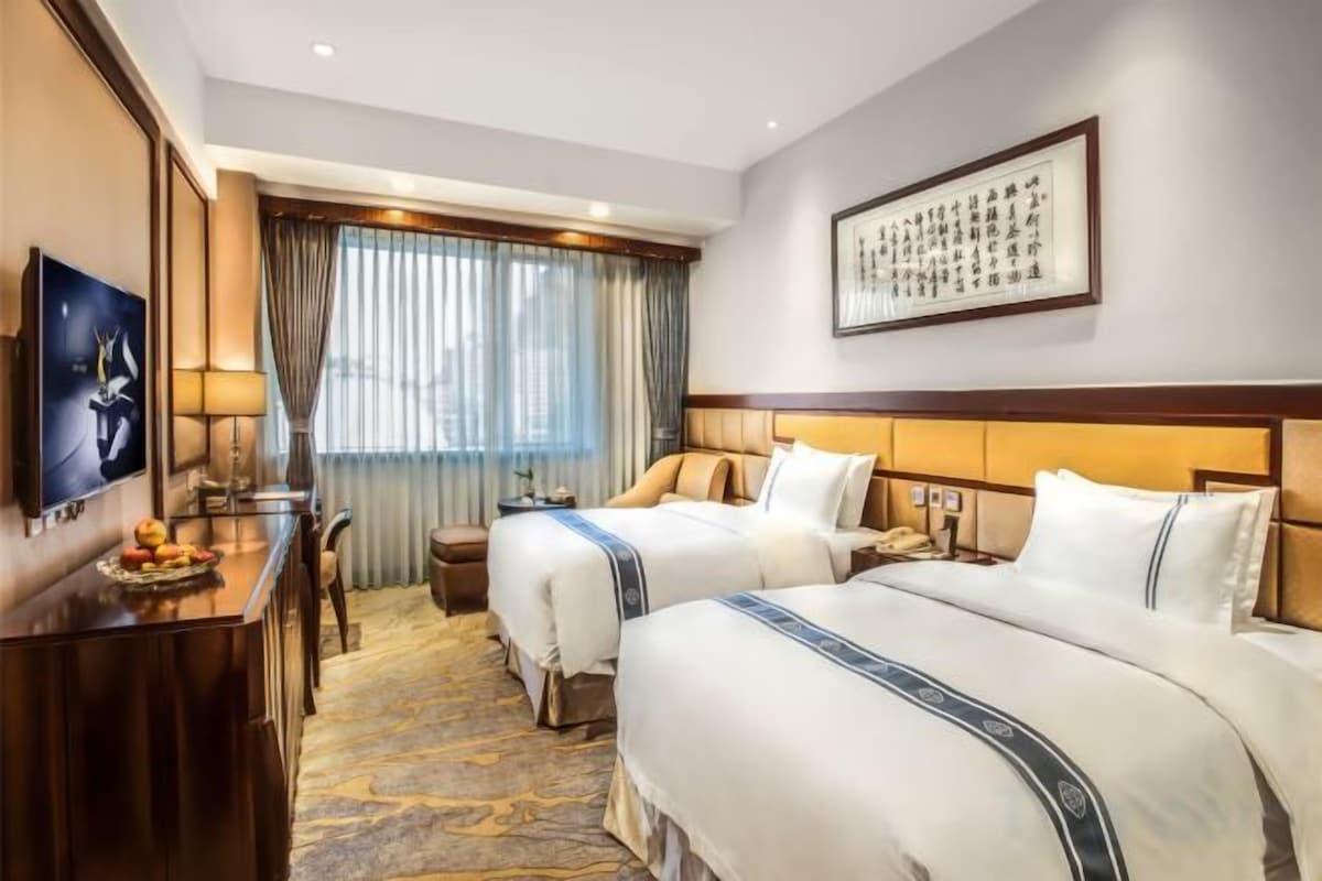 Habitación de hotel con dos camas, un escritorio con silla, un televisor y un amplio ventanal con cortinas.