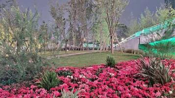 Un jardín arbolado con diversas plantas y flores, un camino y una estructura iluminada en el fondo.