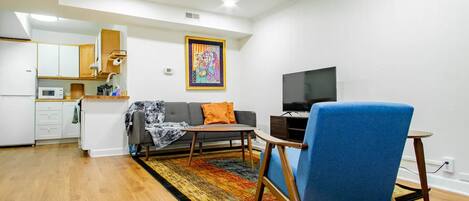 Una sala de estar moderna con un sofá, un sillón azul, una mesa de centro, un televisor y una zona de cocina con armarios y un microondas.