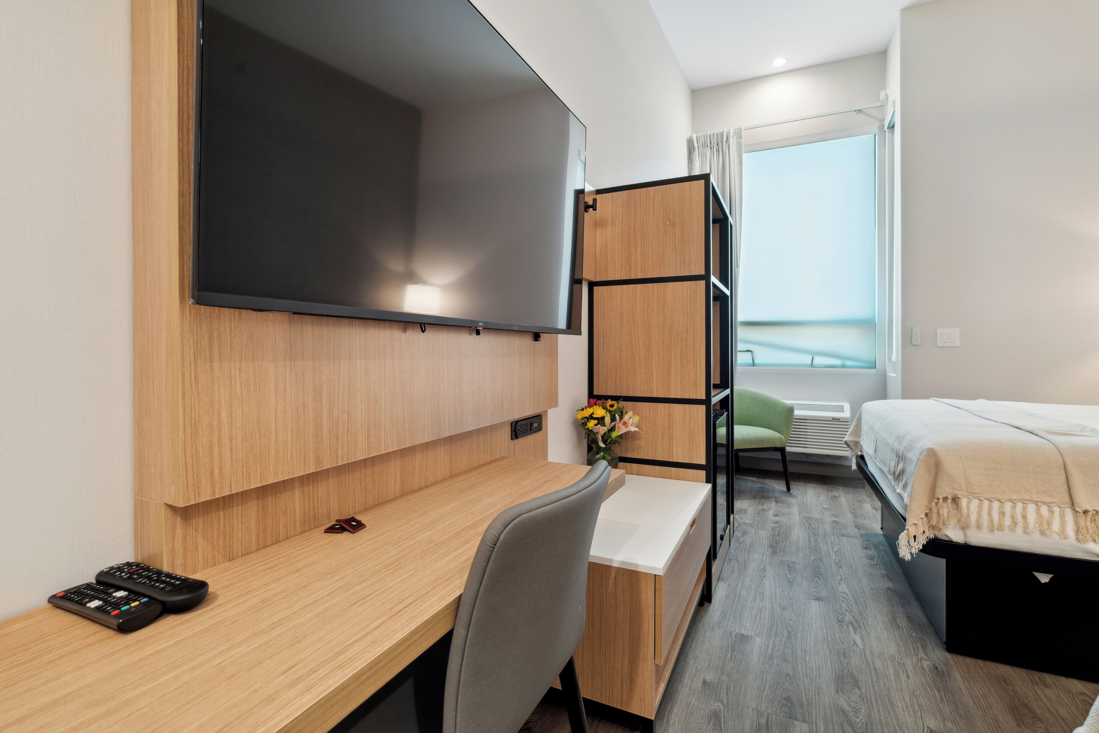 A hotel room with a wooden desk, a bed, a TV mounted on the wall, and a window with a view.