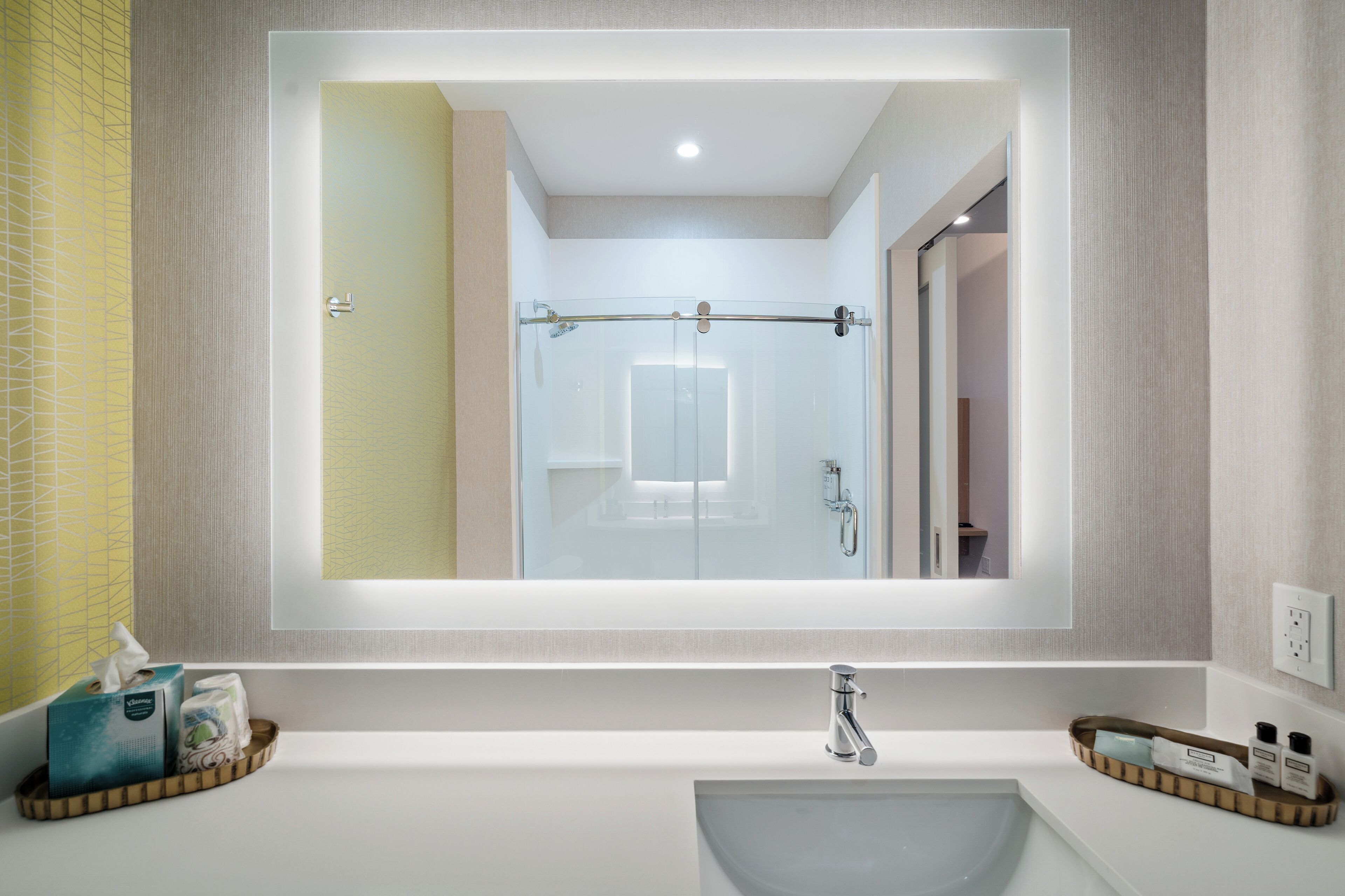 Un baño moderno con un espejo grande, un lavamanos blanco y una cabina de ducha con mampara de vidrio.
