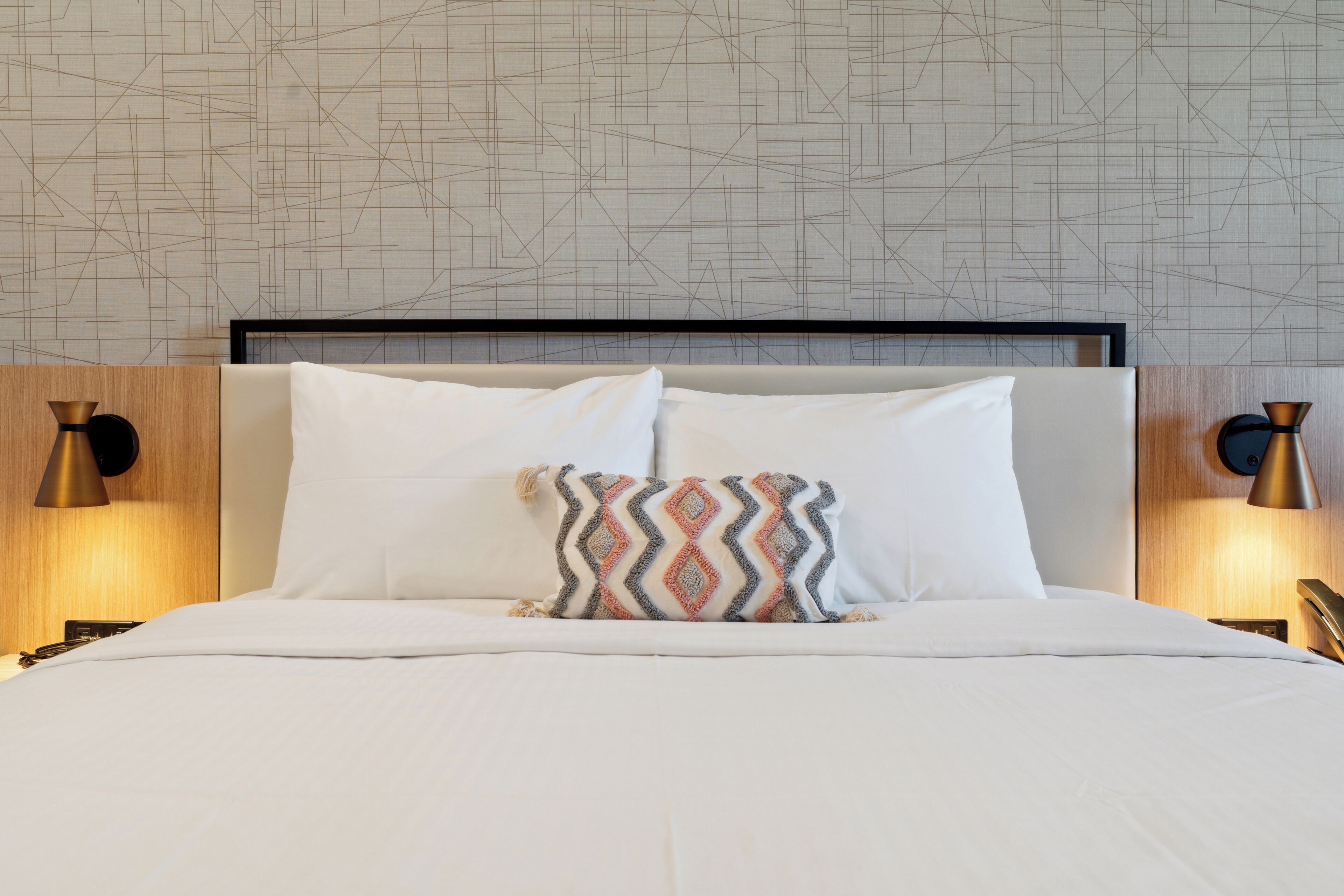 Un lit bien fait, avec du linge de lit blanc et un coussin à motifs, une tête de lit moderne et deux lampes fixées au mur, une de chaque côté.