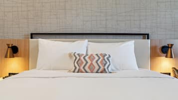 Un lit bien fait, avec du linge de lit blanc et un coussin à motifs, une tête de lit moderne et deux lampes fixées au mur, une de chaque côté.