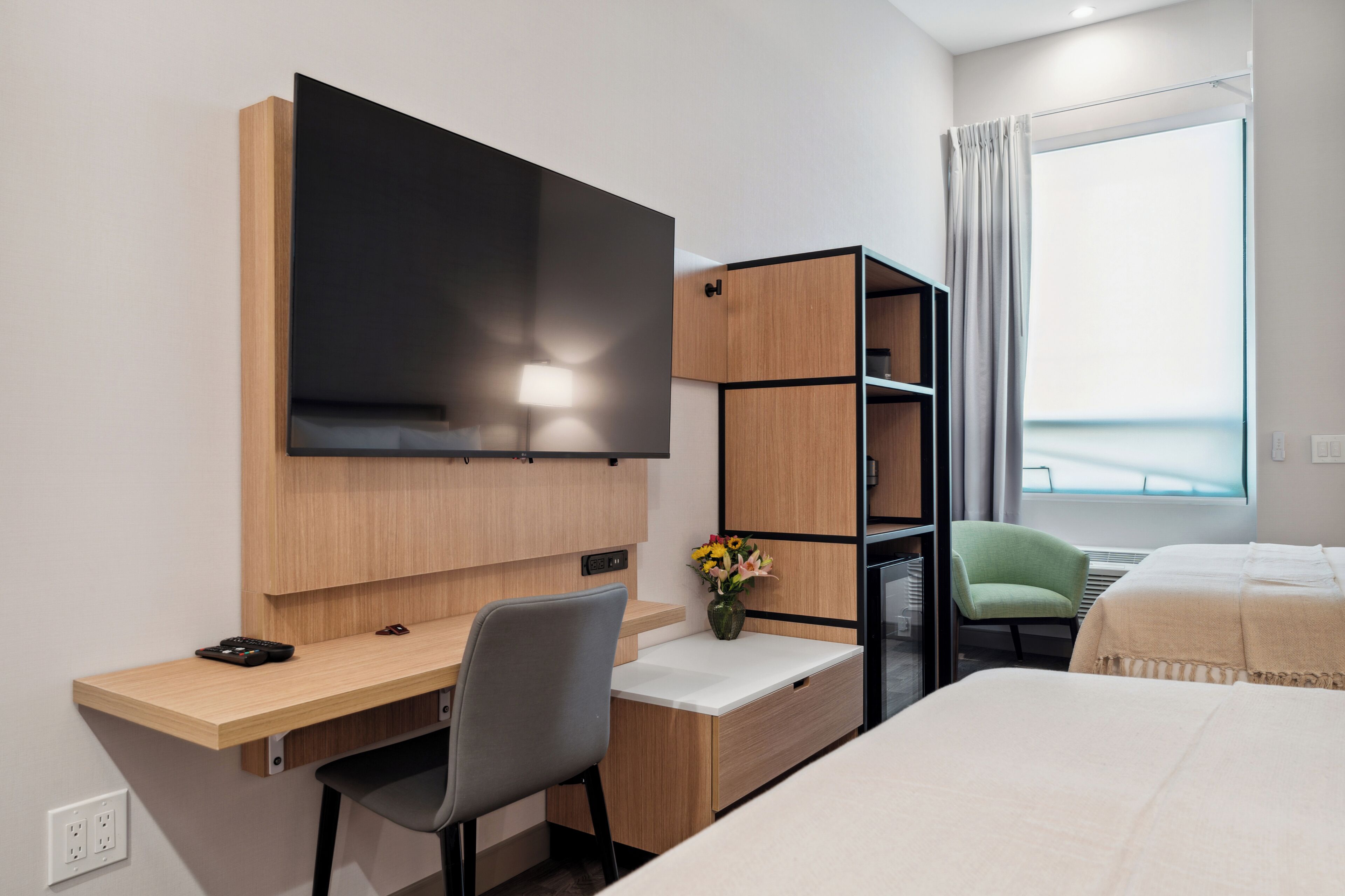 Habitación de hotel con una cama, un escritorio con una silla, un televisor, una mesita con un jarrón de flores y una ventana con cortinas.