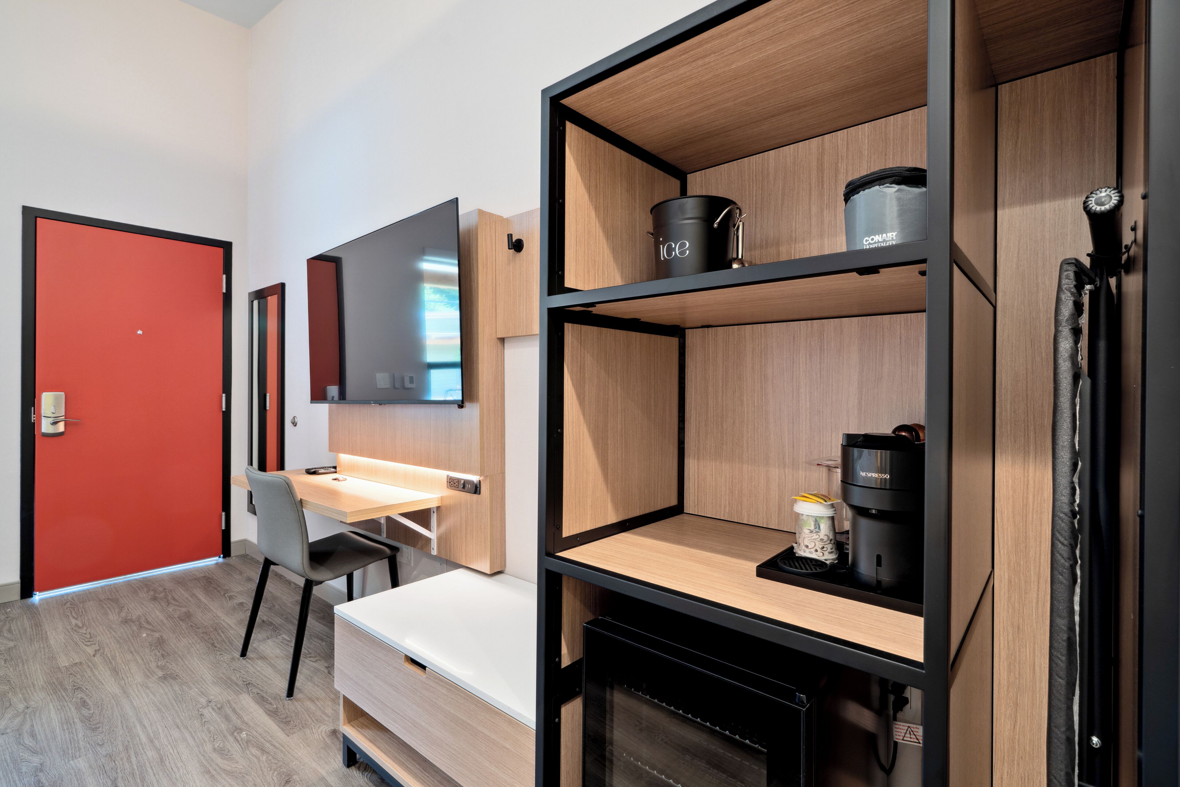 A modern hotel room with a wooden desk, a chair, a bed, a TV, and built-in shelves with a coffee maker and a container labeled "ice".