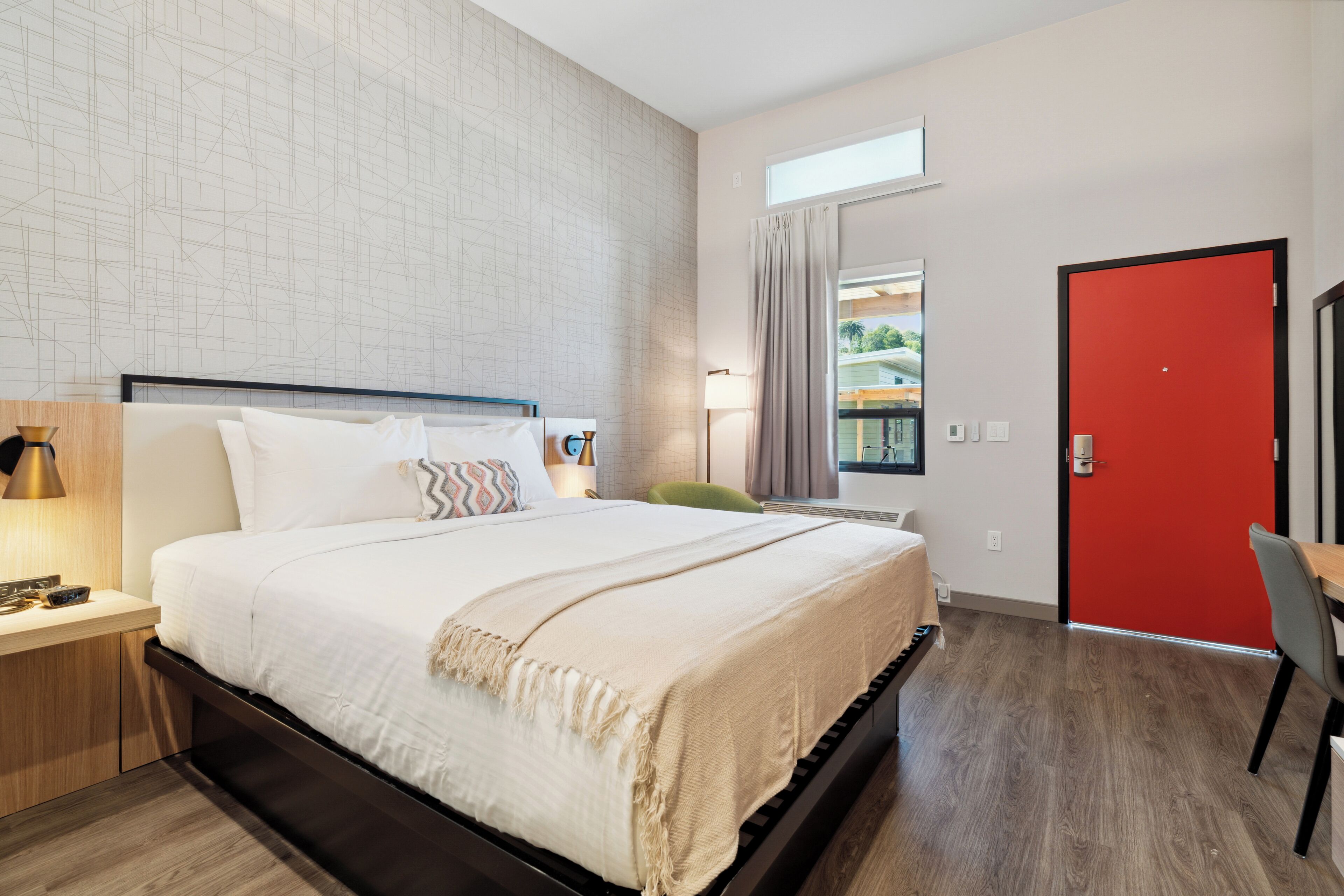 Une chambre d’hôtel moderne avec un grand lit, un bureau, une chaise, une porte rouge et une fenêtre donnant sur l’extérieur.