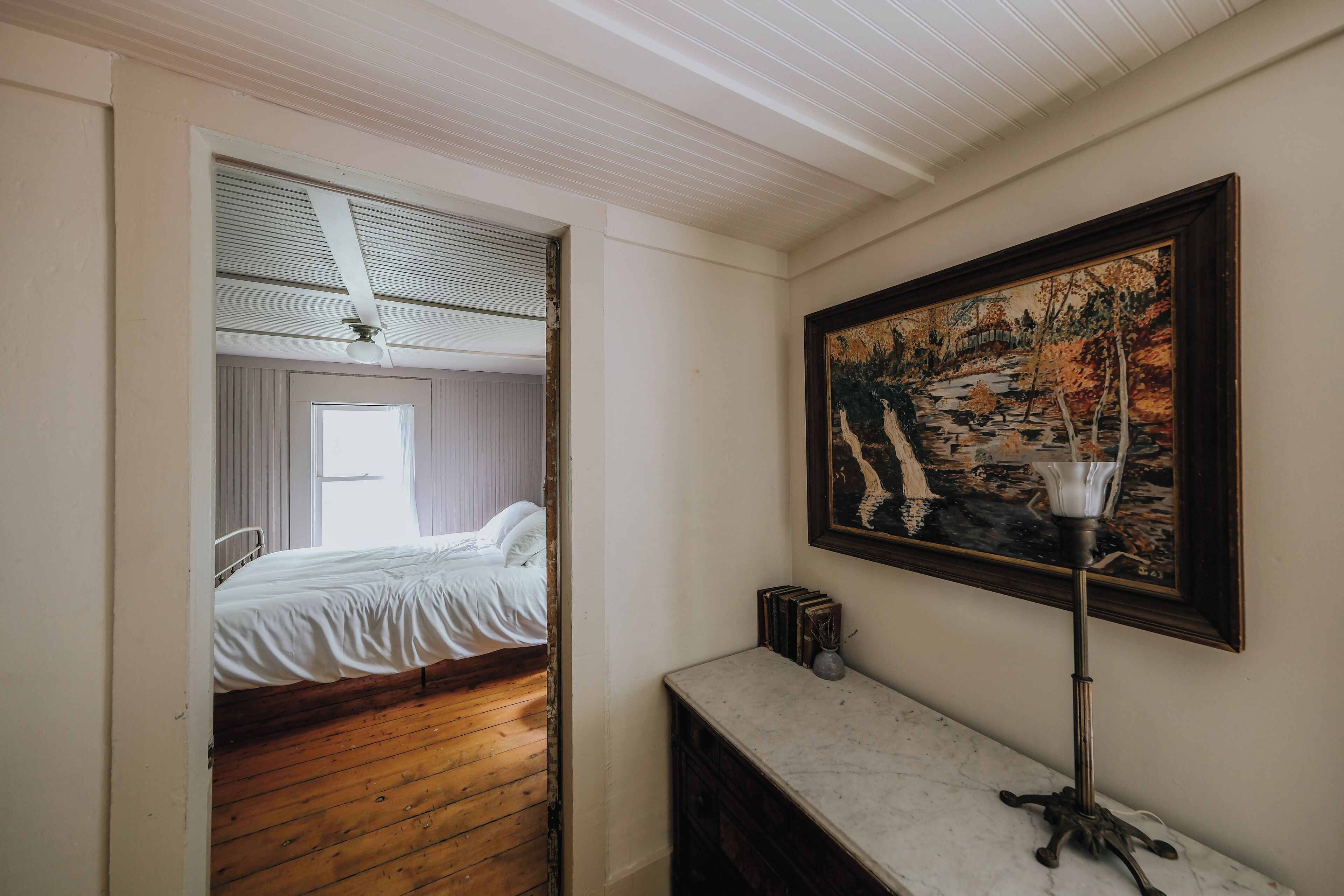 A bedroom with a wooden floor, a bed with white bedding, a window, and a painting on the wall.